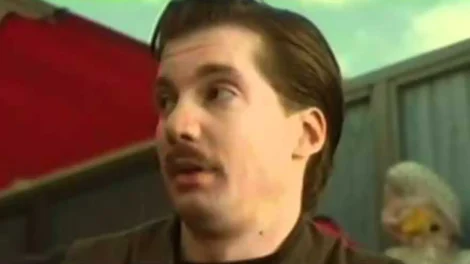 Man with a mustache and slicked-back hair speaking in a scene from a film or video.