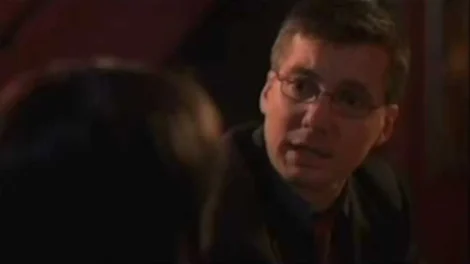 Man wearing a suit and glasses engaged in a conversation with another person in a dimly lit setting.
