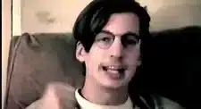 Individual with glasses speaking while seated on a couch, captured in a video format.