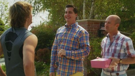 Three men standing outdoors, one holding a pink box, engaged in conversation.