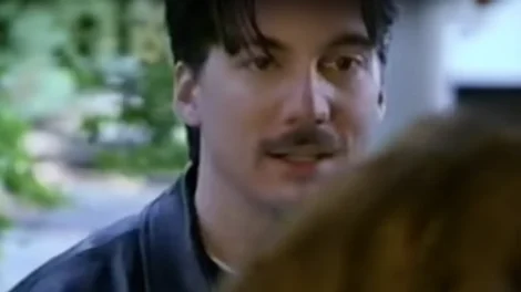 A man with a mustache wearing a dark jacket, engaged in conversation with another person.