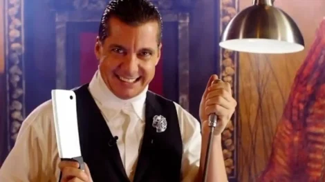 A man in a vest holding a cleaver in one hand and a hook in the other, smiling at the camera.