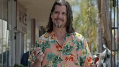 Man with long hair wearing a colorful floral shirt, smiling and holding a small object.