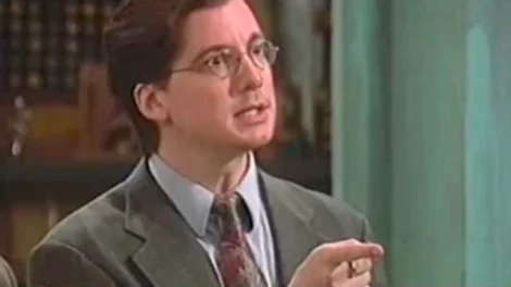 Man wearing a suit and glasses gesturing while speaking in a formal setting.