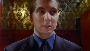 Man wearing a dark suit and blue shirt with a serious expression in a dimly lit setting.