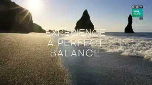 A beach scene featuring a rock formation and waves lapping at the shore under sunlight.