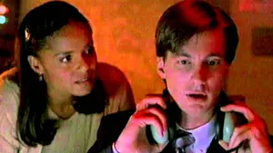 A man wearing headphones looking intently at a computer screen while a woman observes him closely.