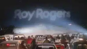 Cars parked at a drive-in movie theater with a large Roy Rogers sign illuminated in the background.