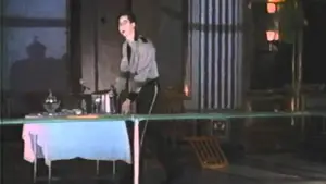 Individual in a gray outfit performing in a dimly lit room with a table and various objects.