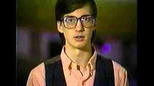 A young person wearing large glasses and a vest, speaking directly to the camera.