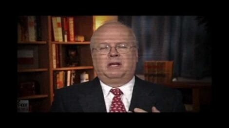 A man with glasses and a suit speaking during a video interview, with bookshelves in the background.