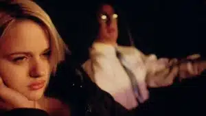 A woman with short blonde hair sitting in a car next to a man wearing glasses and a white shirt.