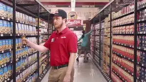 Top 5 Best Commercials Of 2025 | D.c. Douglas Grocery store employee wearing a red shirt and cap standing in an aisle with shelves of products.