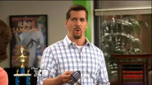 Man in a plaid shirt with a goatee holding a tie in a classroom setting.