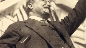 Black and white photograph of a man with glasses and a mustache, raising his hand in a gesture.