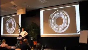 Navigating The Intricacies Of Medical Narration: From Corporate Videos To Patient Education | D.c. Douglas Speaker presenting a mechanical bearing image on a large screen during a conference.