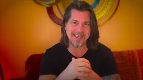 Gadfly: Tweaked News Nipples | D.C. Douglas Man with long hair and a beard smiling while seated in front of colorful wall decorations.
