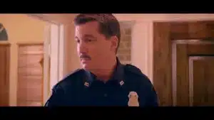 A police officer wearing a uniform with a badge and a mustache, standing indoors.