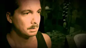 Close-up of a man with a mustache, wearing a black tank top, in a dimly lit environment.