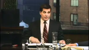 Man wearing a suit and tie seated at a conference table with glassware and papers.