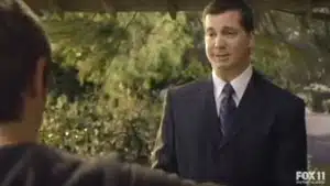 A man in a dark suit and tie speaking to another person outdoors in a casual setting.