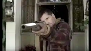 A man aiming a shotgun from a window, wearing a plaid jacket and focused expression.
