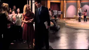 Two television hosts conversing backstage in a studio with crew members visible in the background.