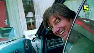 A man with brown hair smiling while leaning out of a car window.