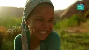 A young girl with a headscarf smiling broadly in a natural setting, showcasing joy.