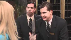 Two male hotel receptionists in black suits and ties engaging with a guest at the front desk.