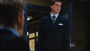 A man in a black suit with a tie standing at a doorway, looking towards another person.