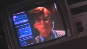 Individual with glasses appearing on a vintage computer monitor displaying data on the screen.
