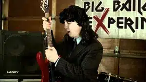 Individual wearing a suit and long black wig holding a red electric guitar while performing.