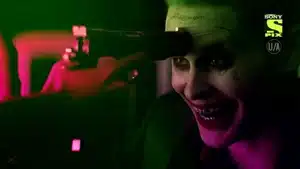 Joker character with green hair and face paint, smiling while a gun is pointed at his forehead.