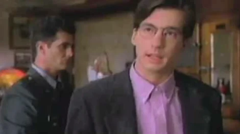 A man wearing glasses and a pink shirt stands in a room with another person in the background.