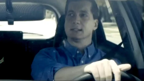 A man with short hair wearing a blue shirt, driving a car while looking ahead.