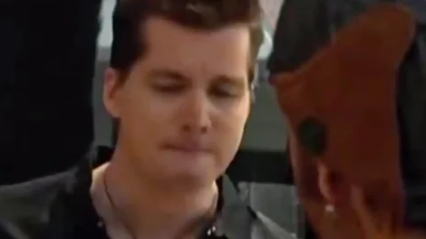 A man with short hair and a neutral expression looking down, wearing a dark shirt.