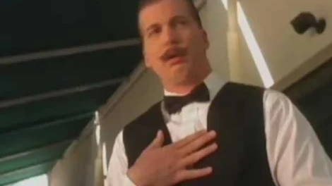 Man wearing a black vest and bow tie with a mustache, standing with a hand on his chest.