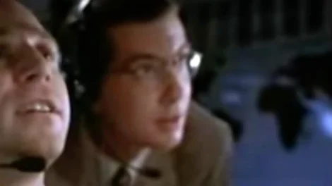 A man wearing glasses and headphones, focused on a task in a dimly lit environment.