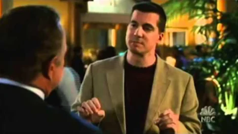 Man wearing a beige blazer and dark shirt speaking to another person in a social setting.