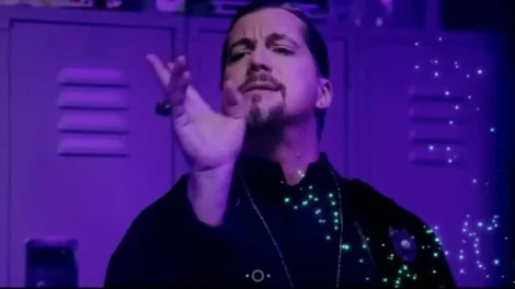 Man wearing a black outfit with a purple background, gesturing with his hand.