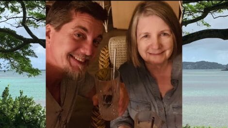 A man and a woman smiling while holding drinks, seated indoors with a tropical backdrop.