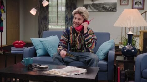 Voice Over: Commercials | D.C. Douglas Individual with light-colored hair talking on a red telephone while seated on a couch in a living room.