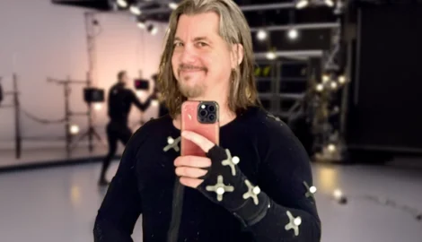 Man wearing a motion capture suit with markers, holding a smartphone for a selfie in a studio setting.